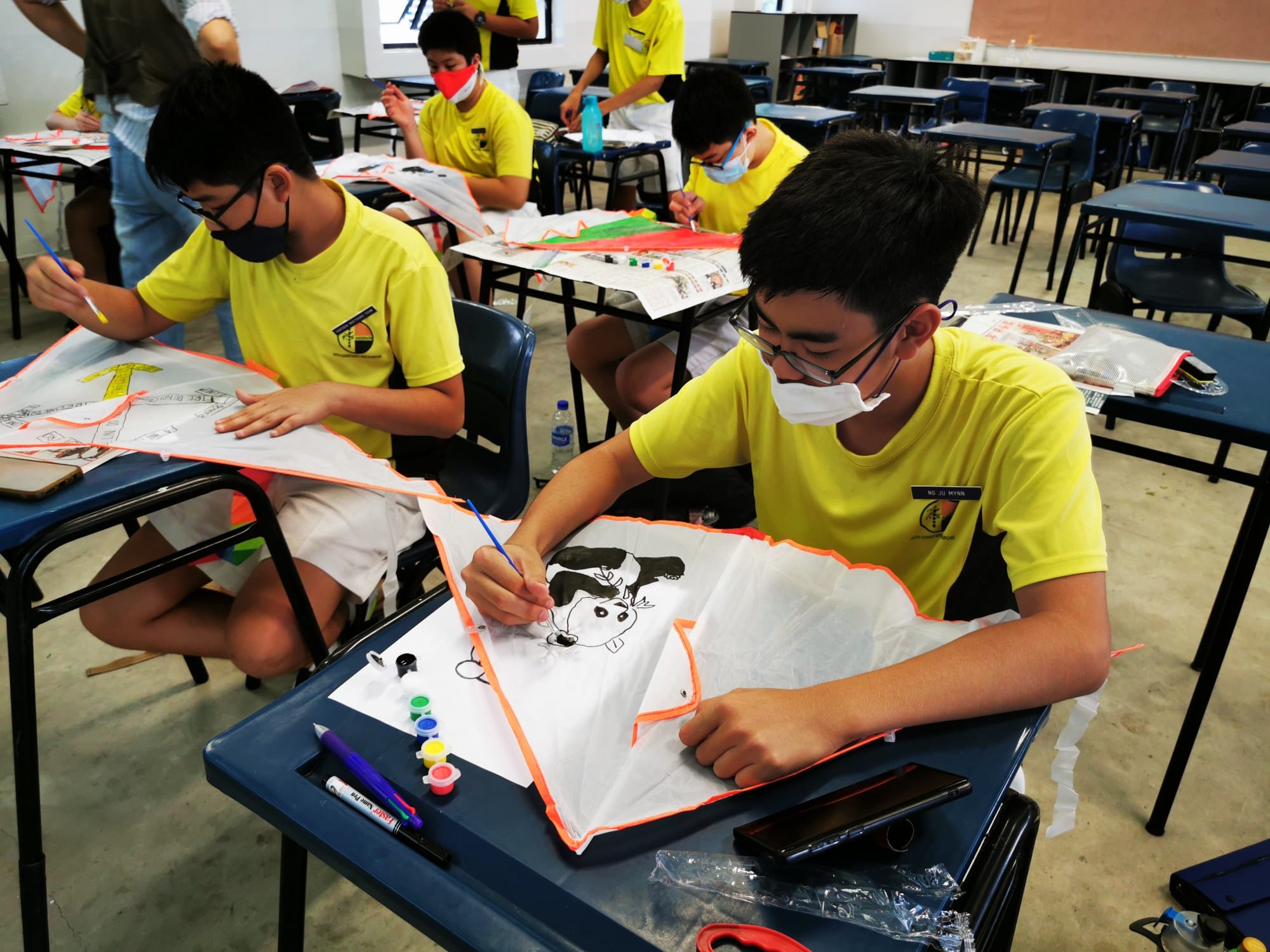 Students in yellow shirts and masks paint designs on white fabric kites at school desks in a classroom.